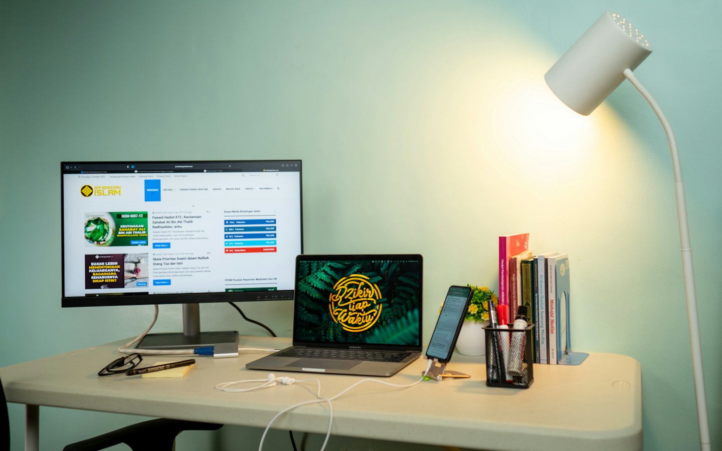 Uspořádaný stůl s notebookem, monitorem, telefonem a lampou.